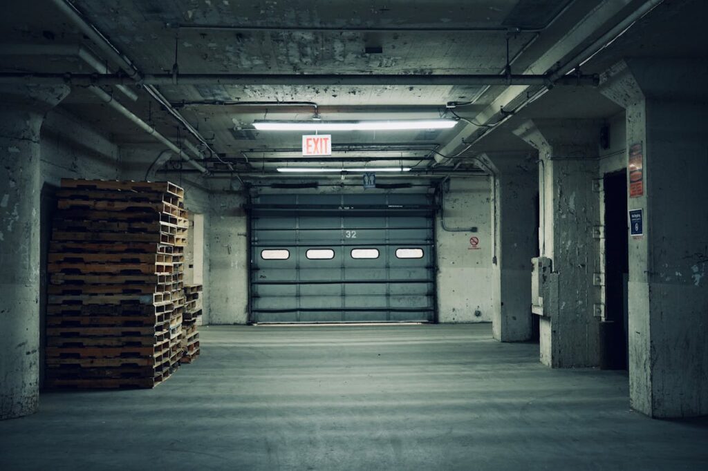 Empty industrial warehouse featuring stacked pallets and a closed garage door under fluorescent lighting.